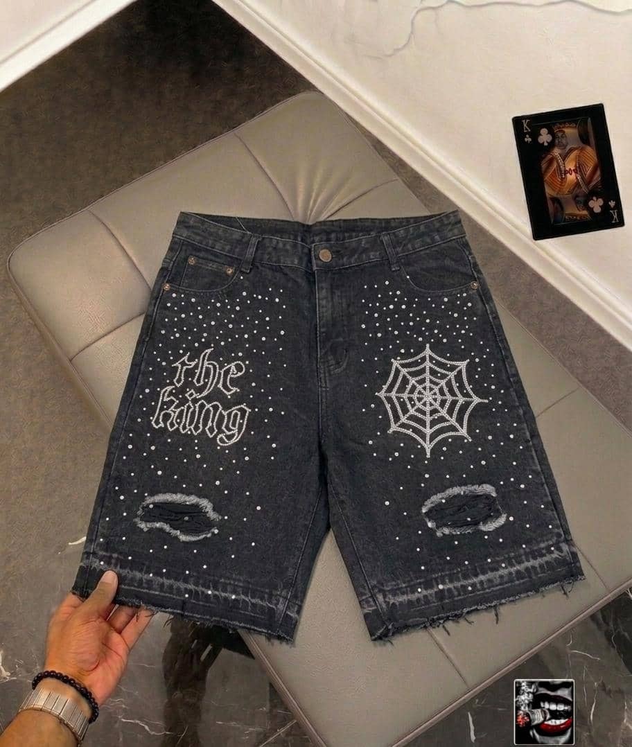 Front view of dark denim jorts with white rhinestone 'the king' text, spider web and dot patterns, plus distressed rips.