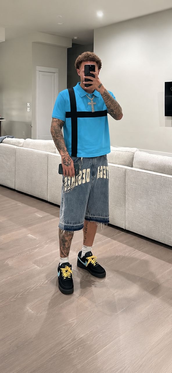 Man wearing blue polo shirt with black cross design, styled with denim shorts and sneakers.