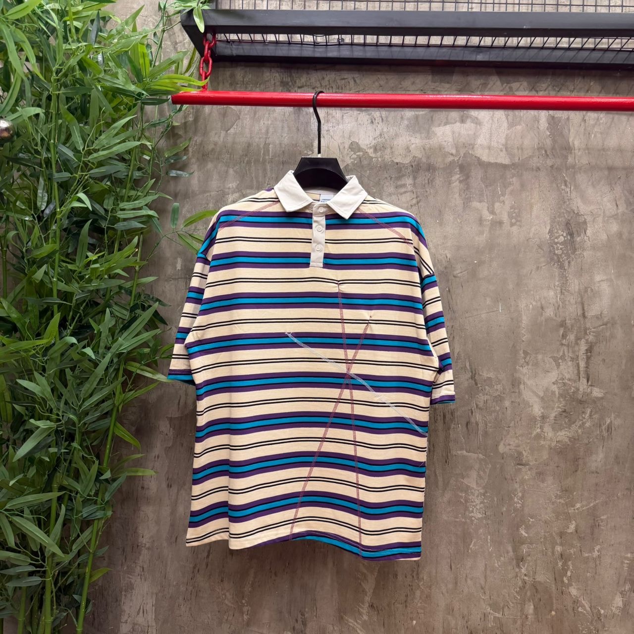 Cream striped polo with purple teal and black stripes plus contrast stitching, hanging on red rack with plants.