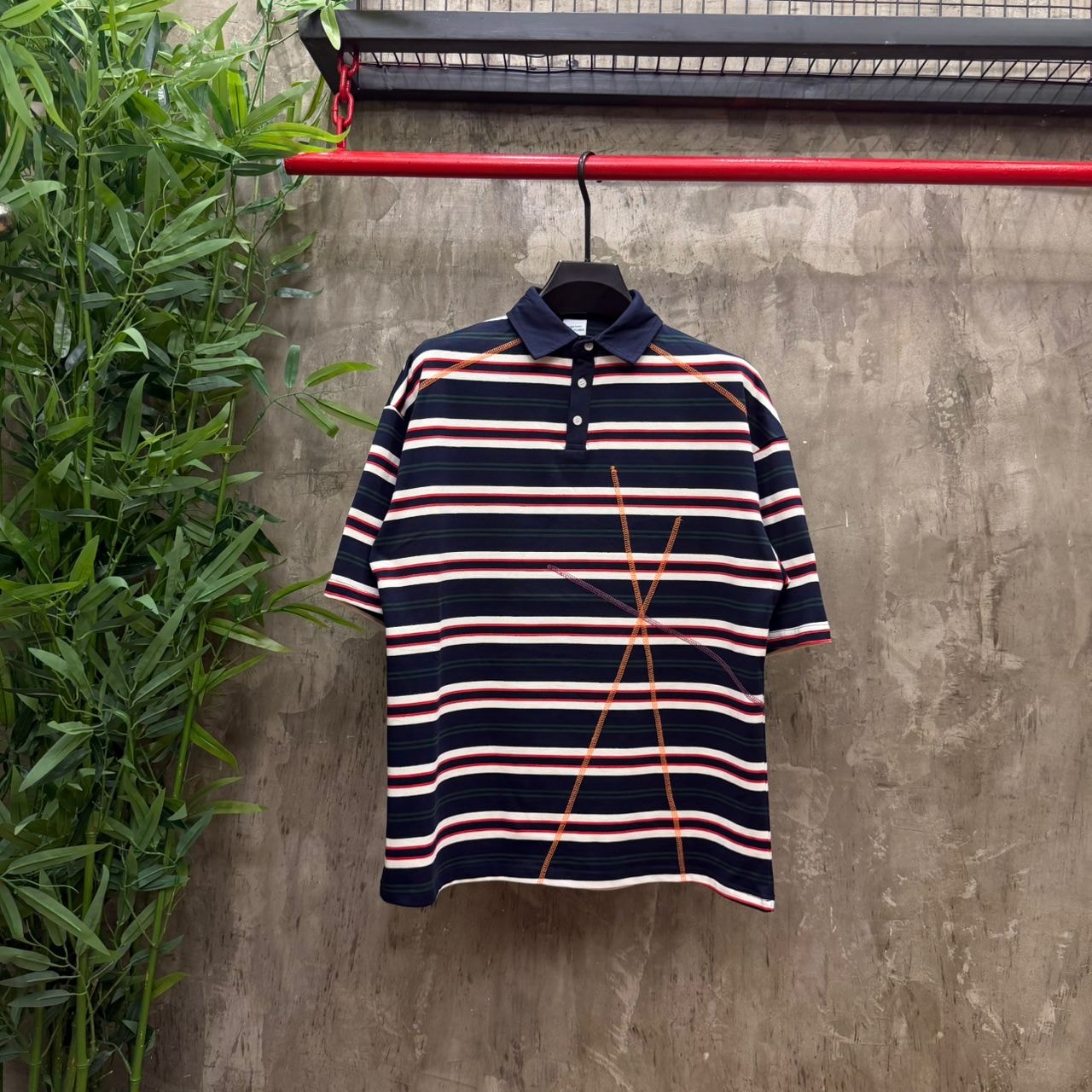 Navy striped polo with orange contrast stitching hanging on red rack with plants in background.