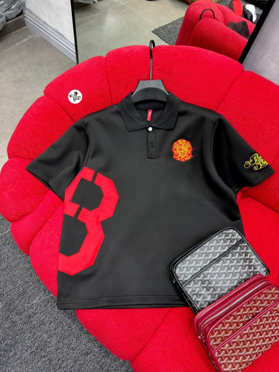 Black polo shirt with large red number 8 print on front, red crest on chest and yellow embroidery on sleeve, displayed on red couch.