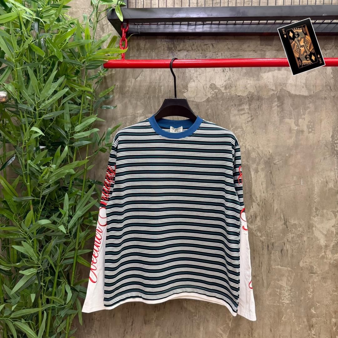 Front view of green and white striped long sleeve t-shirt hanging against textured wall with plants.