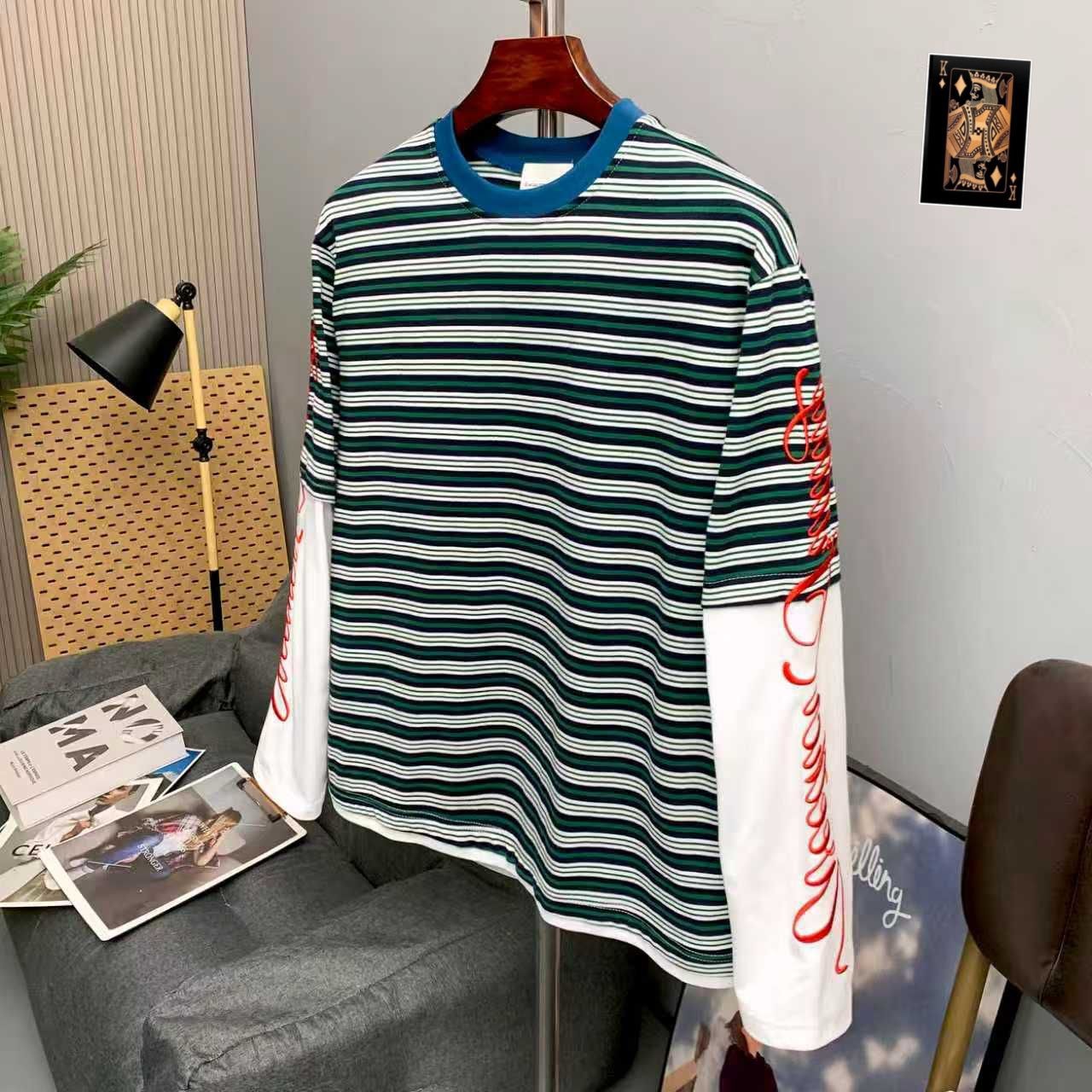 Green and white striped long sleeve t-shirt on wooden hanger in modern interior, showing red embroidery on white sleeve.