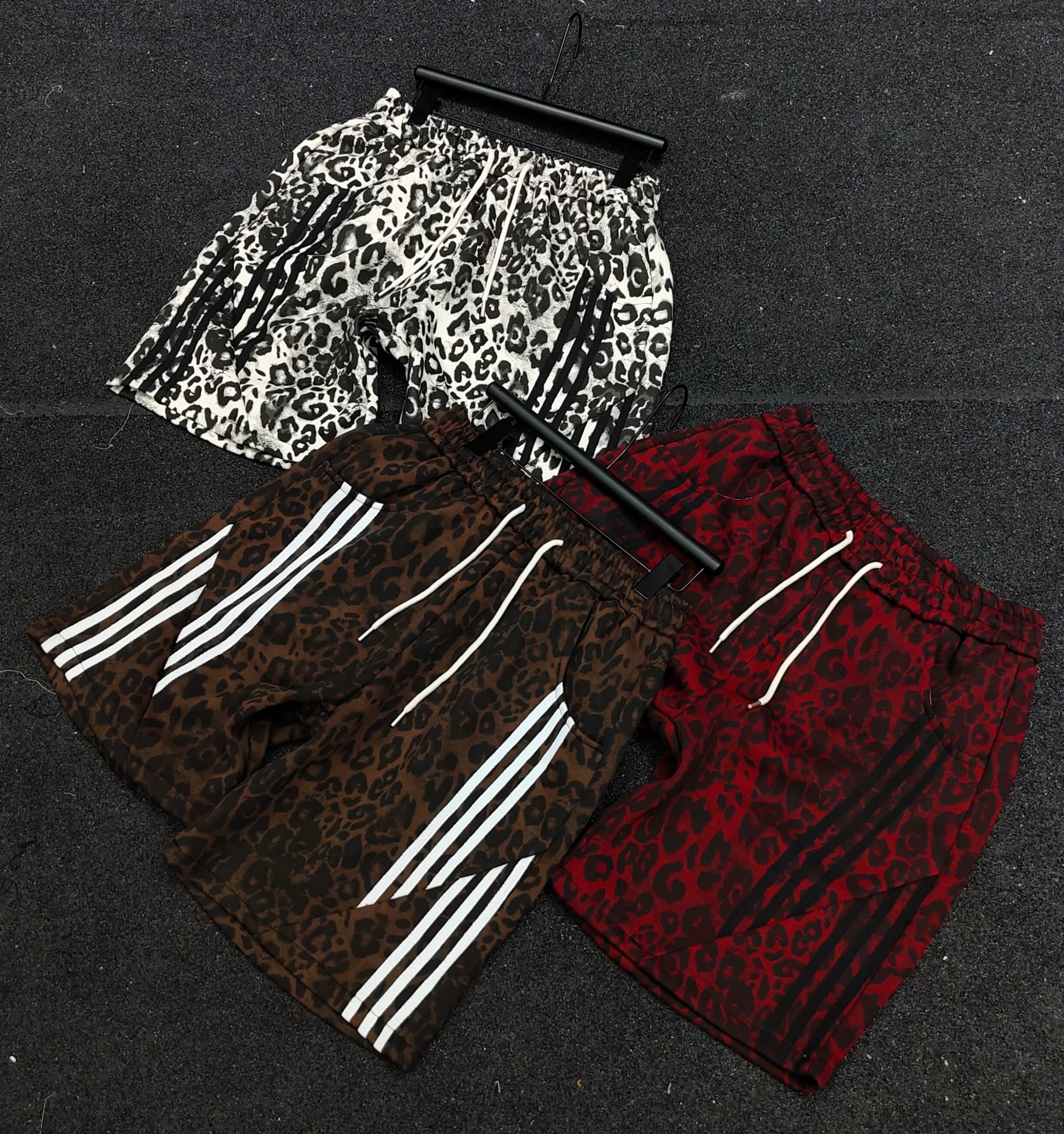 Three leopard print shorts in White/Black, Brown, and Red with white side stripes and drawstrings laid on dark pavement.