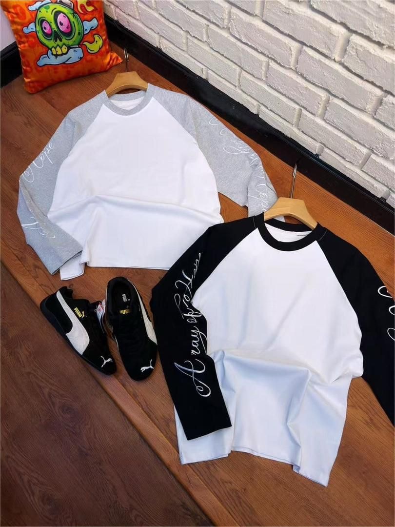 Two raglan long sleeve shirts with white bodies and gray or black contrasting sleeves with cursive prints, displayed with sneakers on wooden floor.
