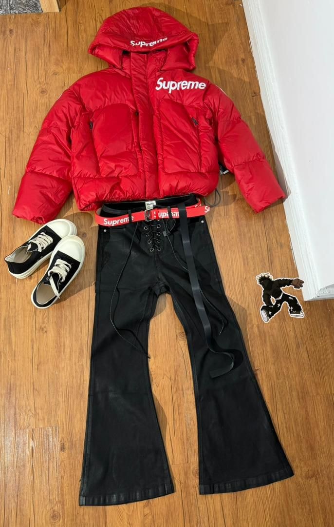 Supreme red puffer jacket styled as full outfit with black pants, red belt, and sneakers on wooden floor.
