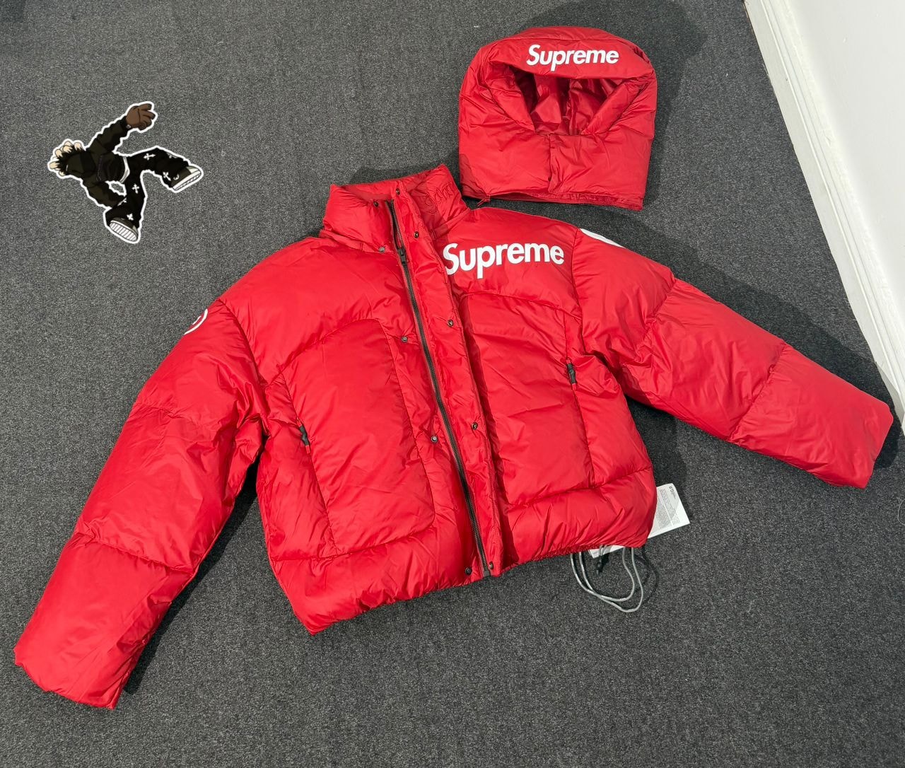 Red Supreme puffer jacket laid flat on gray surface with detached hood and graphic sticker.