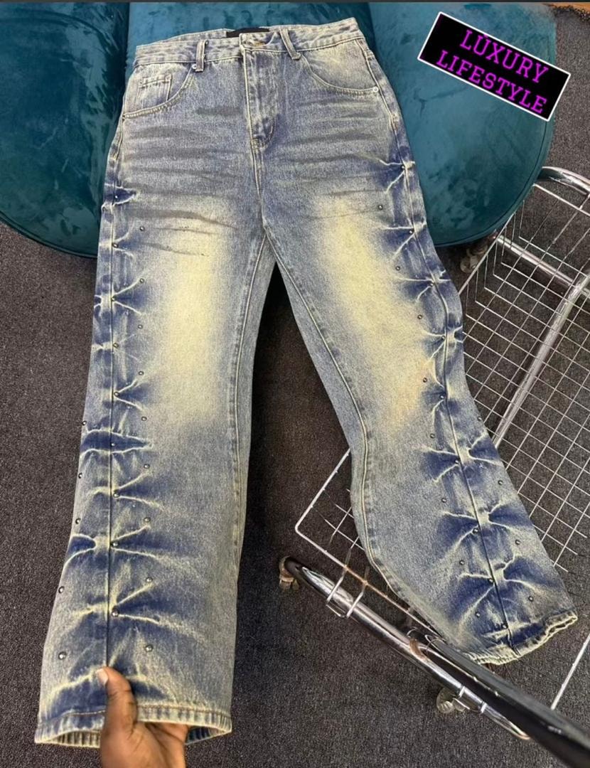 Top down view of bleached blue studded baggy jeans with hand holding the hem on a dark floor.